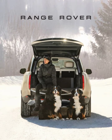 A woman and two Bernese Mountain Dogs pose in front of a Range Rover with its trunk open in a snowy, mountainous landscape.