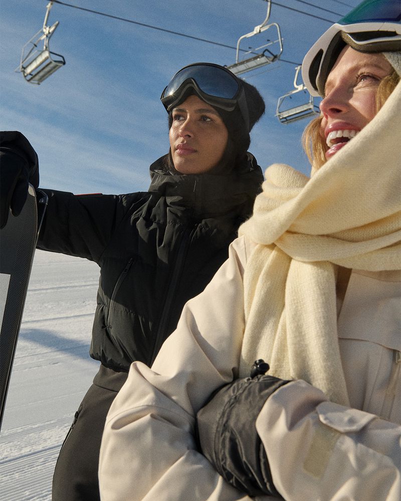 Two women in winter gear stand in a snowy mountain landscape. One wears goggles and a black puffer jacket, the other a white scarf.