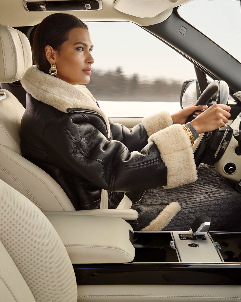 Adult female driving a luxury sport utility vehicle (SUV) on a snowy road, wearing a stylish black leather jacket with fur collar.