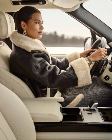 Adult female driving a luxury sport utility vehicle (SUV) on a snowy road, wearing a stylish black leather jacket with fur collar.