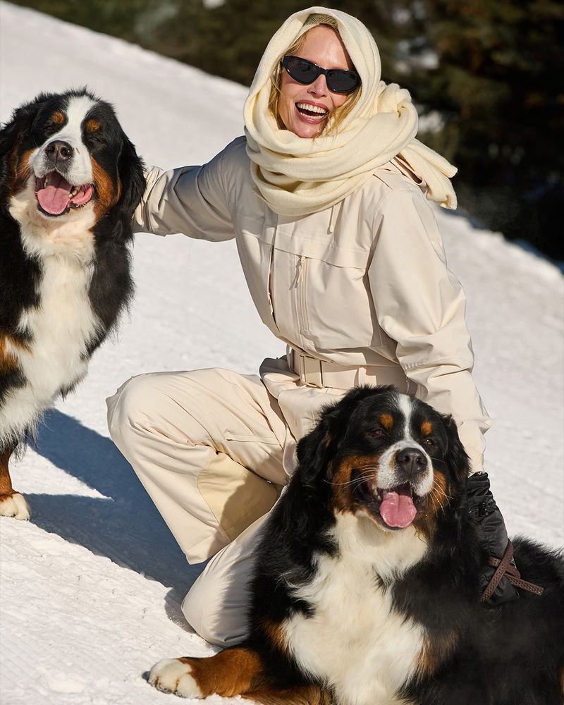 Woman in cream ski suit kneeling in snow with two Bernese Mountain Dogs, smiling, wearing sunglasses and headscarf, outdoor winter scene.