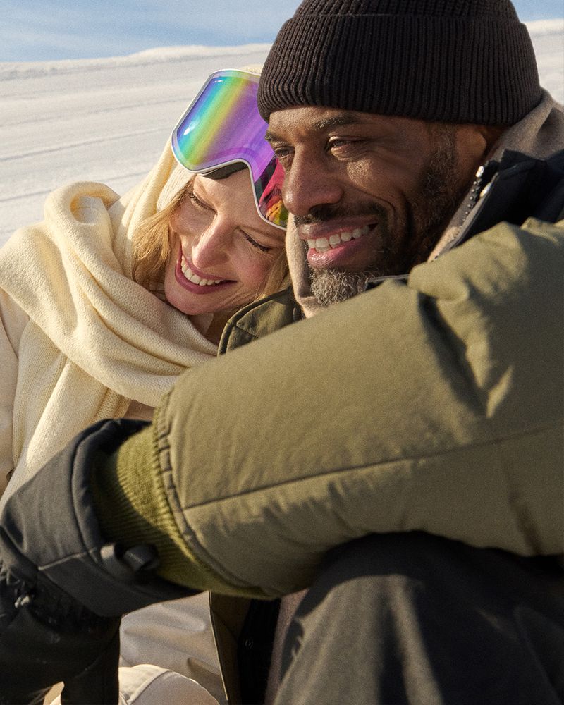 Smiling couple wearing winter outerwear and goggles embrace against a snowy backdrop on a bright day, capturing a moment of warmth.