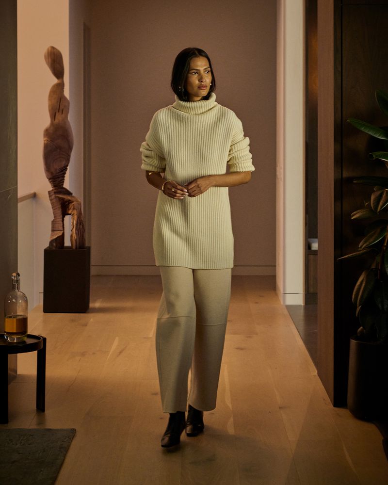Indoor portrait of a woman in a cream-colored sweater and trousers, walking down a hallway with modern decor and natural light.