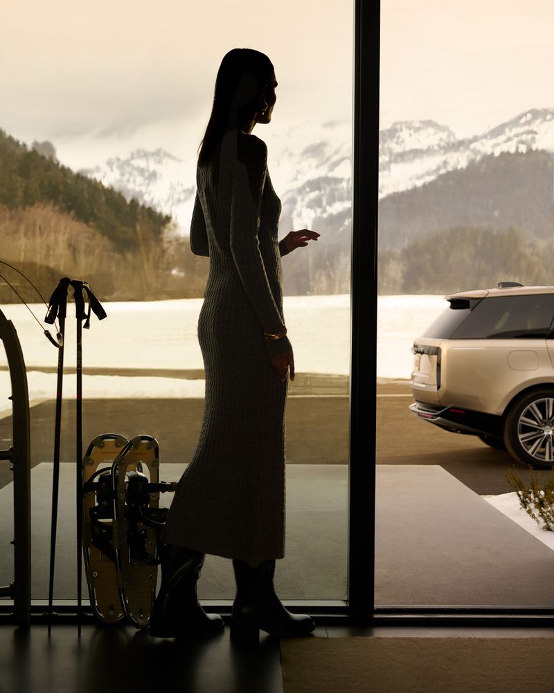 Woman in dress looks out at snowy mountains. Car parked outside the door. Skis and snowshoes indoors.
