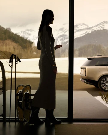 Woman in dress looks out at snowy mountains. Car parked outside the door. Skis and snowshoes indoors.