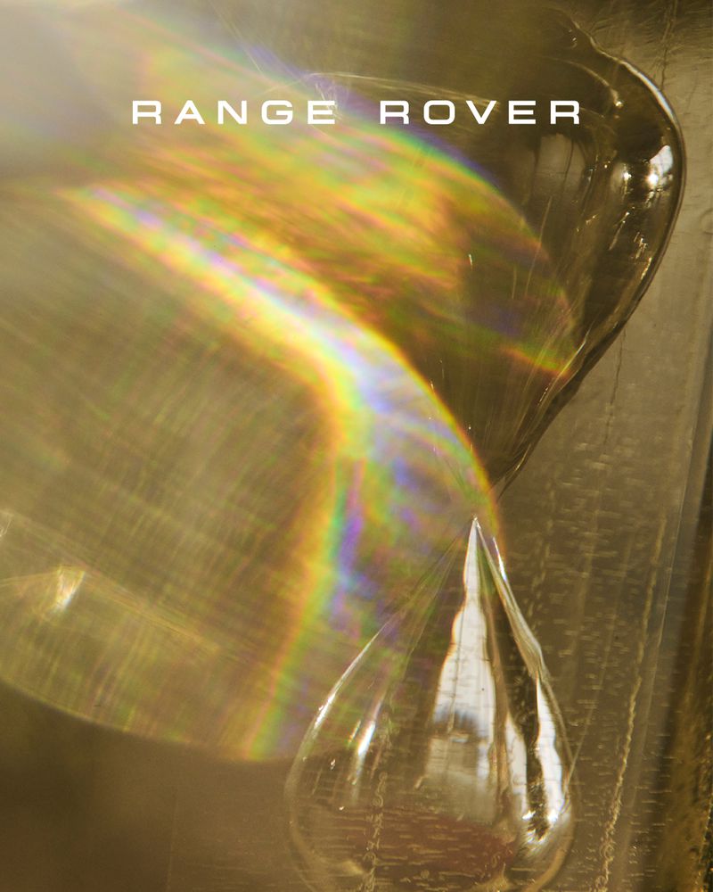 Artistic shot of Range Rover logo with water drop, light refraction, and abstract textures in a warm, golden color palette.