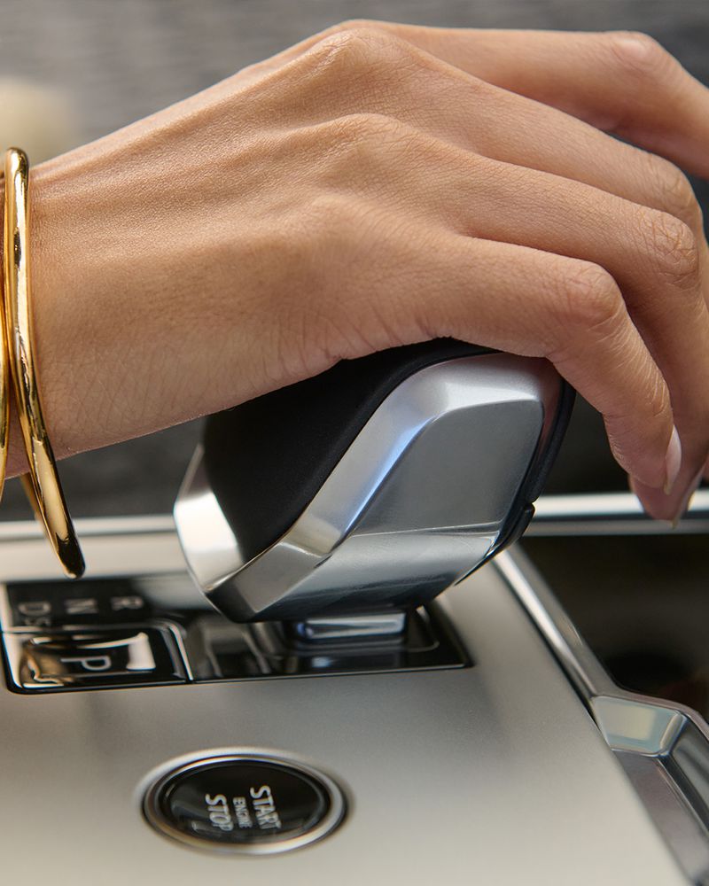 Close-up of a woman's hand with a gold bracelet operating the gear shift in a car, showcasing the vehicle's interior and design.