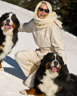 Woman in a cream ski jumpsuit and sunglasses smiling while crouching in the snow with two Bernese Mountain Dogs.