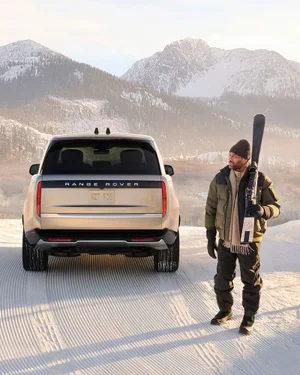 Man holding skis standing on snow next to a champagne gold Range Rover SUV against a snowy mountain background.