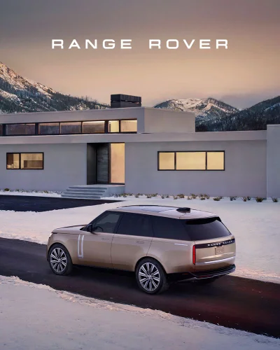 A champagne Range Rover SUV parked on a snowy driveway outside a modern minimalist house with mountains in the background.