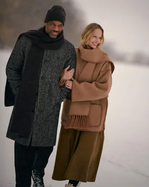 Man and woman in winter coats walking arm in arm across a snowy landscape.