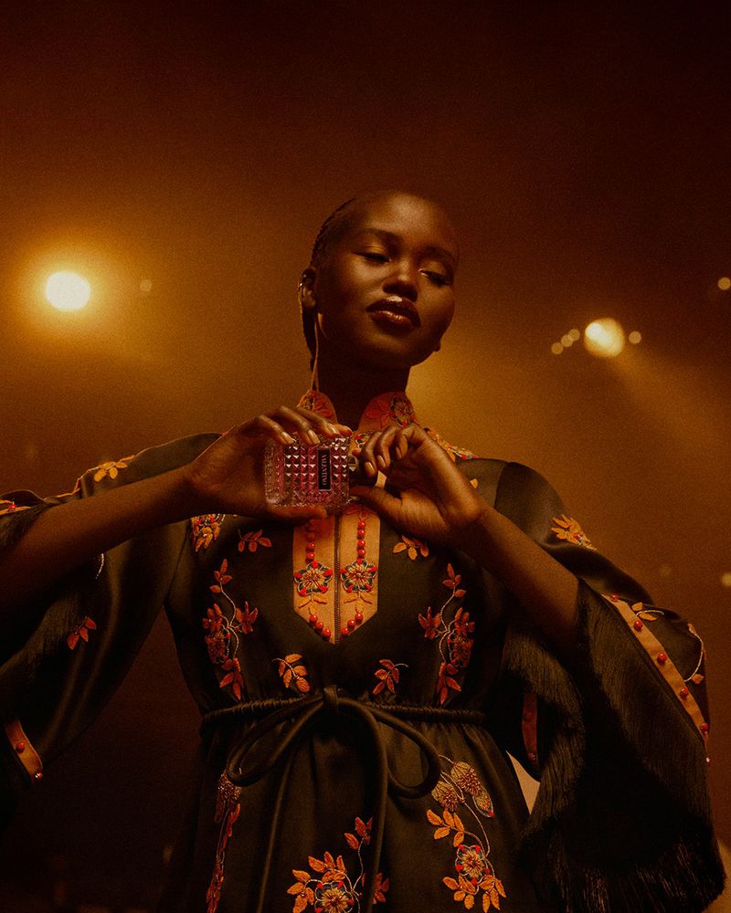 Woman with dark skin poses holding a perfume bottle in warm, golden light, wearing an embroidered, dark green robe.