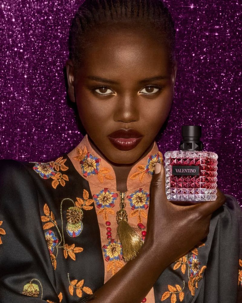 Dark-skinned model poses indoors with Valentino Born in Roma Donna perfume against a glittery purple backdrop, wearing patterned top.