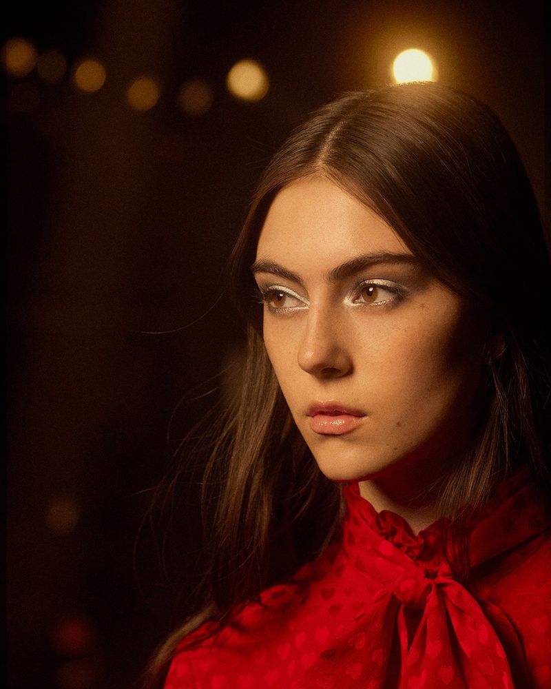 Close-up portrait of a young woman with brown hair and white eyeshadow, wearing a red blouse with bow, against a dark background.