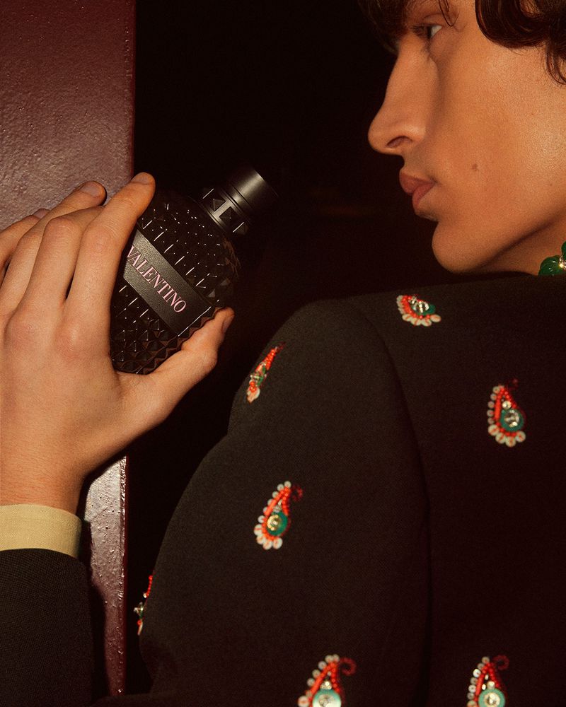 Man holding Valentino cologne bottle. He is wearing an embellished black jacket with paisley pattern in dark indoor environment.