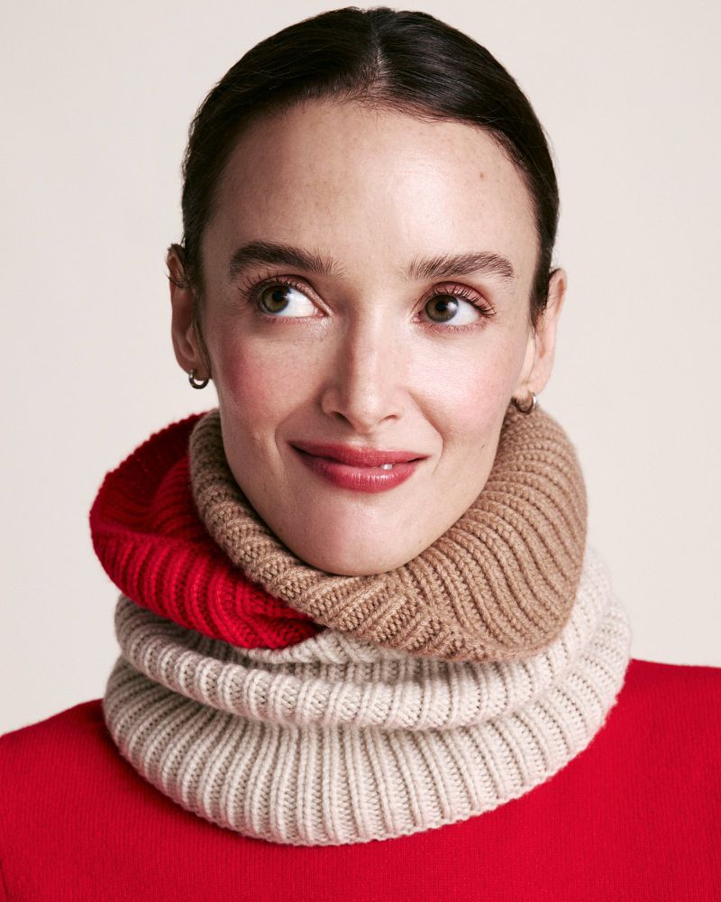 Close-up of a smiling woman wearing a colorful knitted cowl neck warmer in red, beige, and white, against a light background.