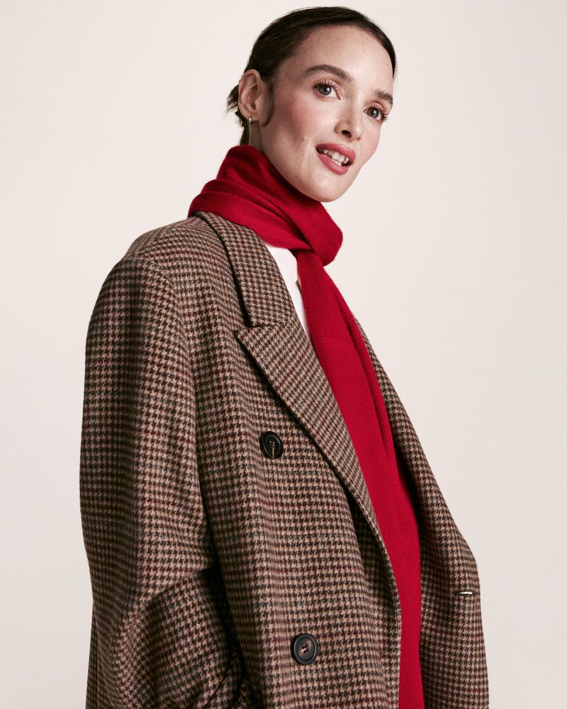 A woman wearing a patterned brown blazer and a red scarf is posing in front of a light-colored background in a fashion portrait.