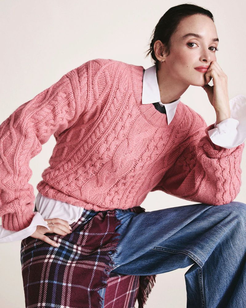 Adult female wearing a pink cable-knit sweater, blue jeans, and a plaid skirt panel poses in a studio with soft lighting.