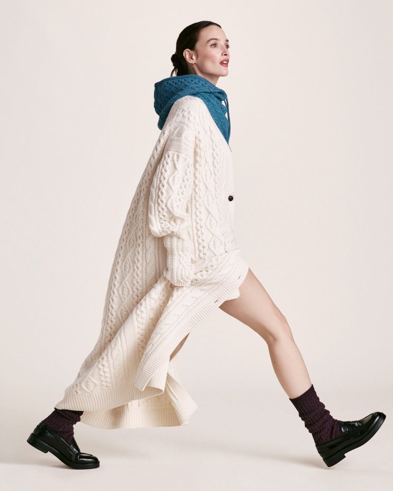 A woman strides confidently in a fashionable ensemble: oversized knit cardigan, stylish scarf-hood, dark socks, and glossy black loafers.