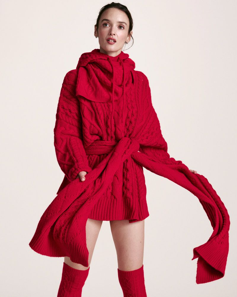 A woman in a red knitted hooded sweater, posing in front of a neutral background, accessorized with matching knee-high socks.