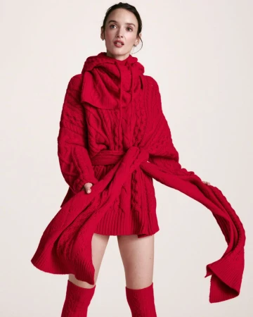A woman in a red knitted hooded sweater, posing in front of a neutral background, accessorized with matching knee-high socks.