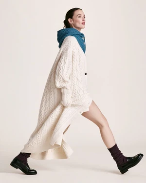 Charlotte Le Bon strides in an oversized cream cable-knit cardigan, teal knit hood, and black loafers against a studio backdrop.