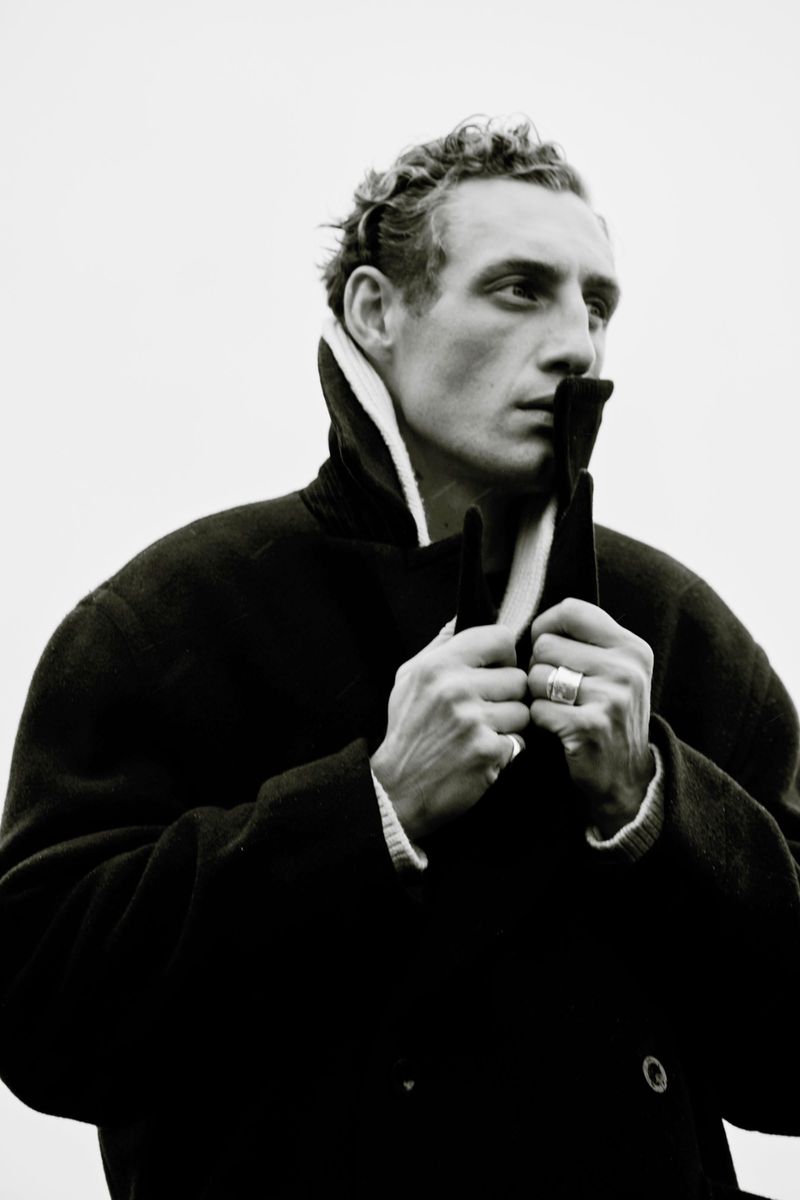Monochromatic portrait of an adult male with curly hair, wearing a dark coat with a contrasting collar, looking off to the side.