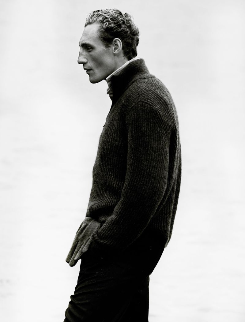 Monochromatic portrait of a slender adult male in a zip-up sweater, hands in pockets, captured in profile with a blurred background.