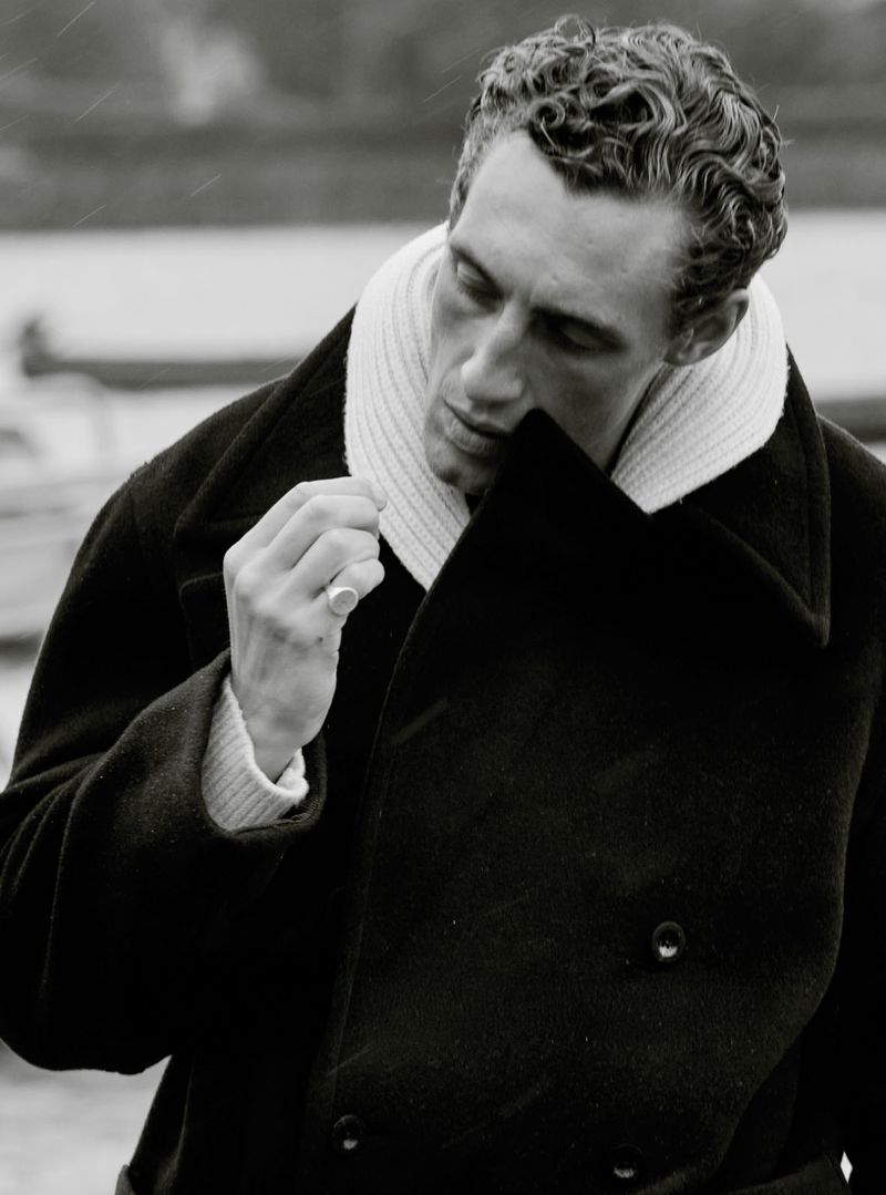 Black and white portrait of adult male wearing dark overcoat with sweater, ring on left hand, outdoors with blurred background.
