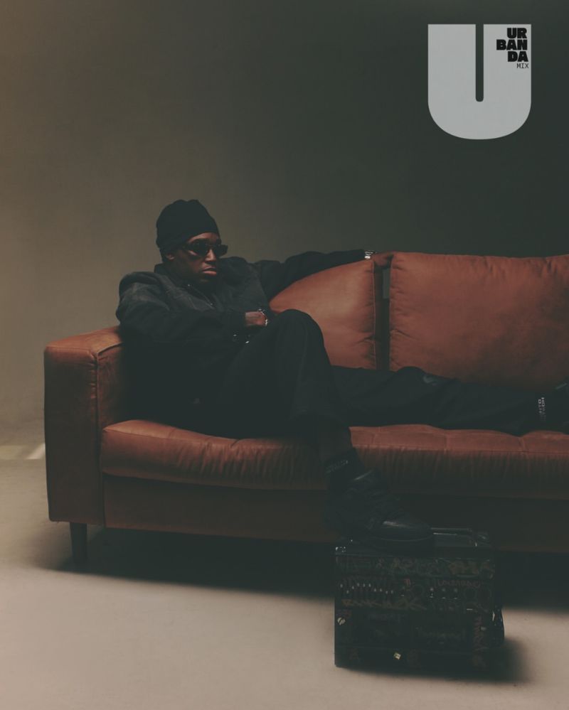 Musician Sech wearing black sunglasses and a beanie while lounging across a brown leather couch in a moody studio setting.