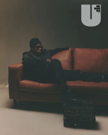 Musician Sech wearing black sunglasses and a beanie while lounging across a brown leather couch in a moody studio setting.