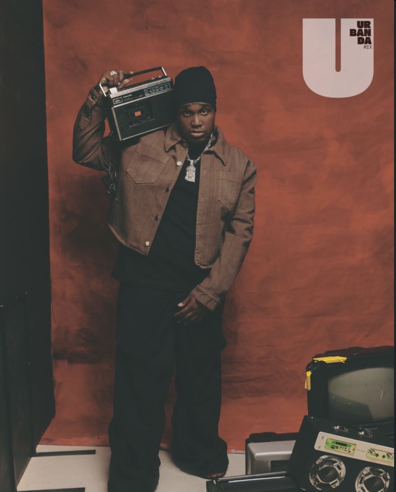 Portrait of an adult male in a jacket and beanie, carrying a boombox, with a television and electronics in the foreground.