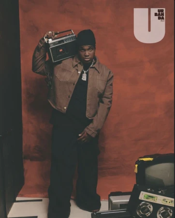 Portrait of an adult male in a jacket and beanie, carrying a boombox, with a television and electronics in the foreground.