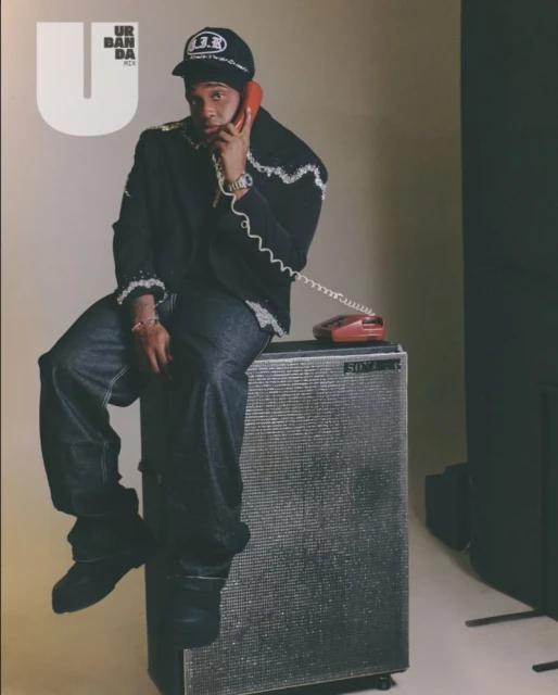 Man in jeans and baseball cap sits on a speaker while talking on a red telephone, wearing a black blazer with white embroidery.