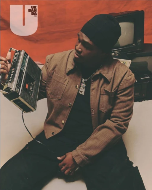 Man in beanie holds vintage tape recorder in front of a faded orange backdrop with an old television set and magazine cover visible.
