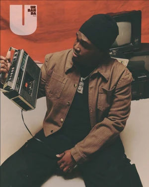 Sech seated on a studio floor holding a vintage cassette recorder, wearing a brown jacket and black beanie.