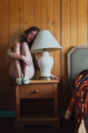 An adult woman in a gold dress is sitting on a bedside table next to a vintage lamp in a wood-paneled bedroom with a colorful quilt.