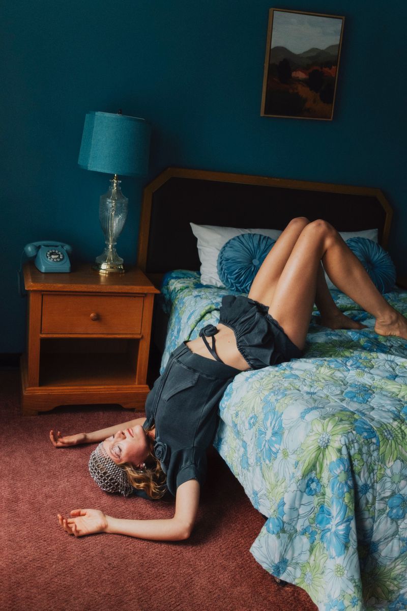 Woman lying on a bed in a blue room with vintage decor, featuring a rotary phone, landscape painting, and floral bedspread.