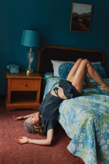 Woman lying on a bed in a blue room with vintage decor, featuring a rotary phone, landscape painting, and floral bedspread.
