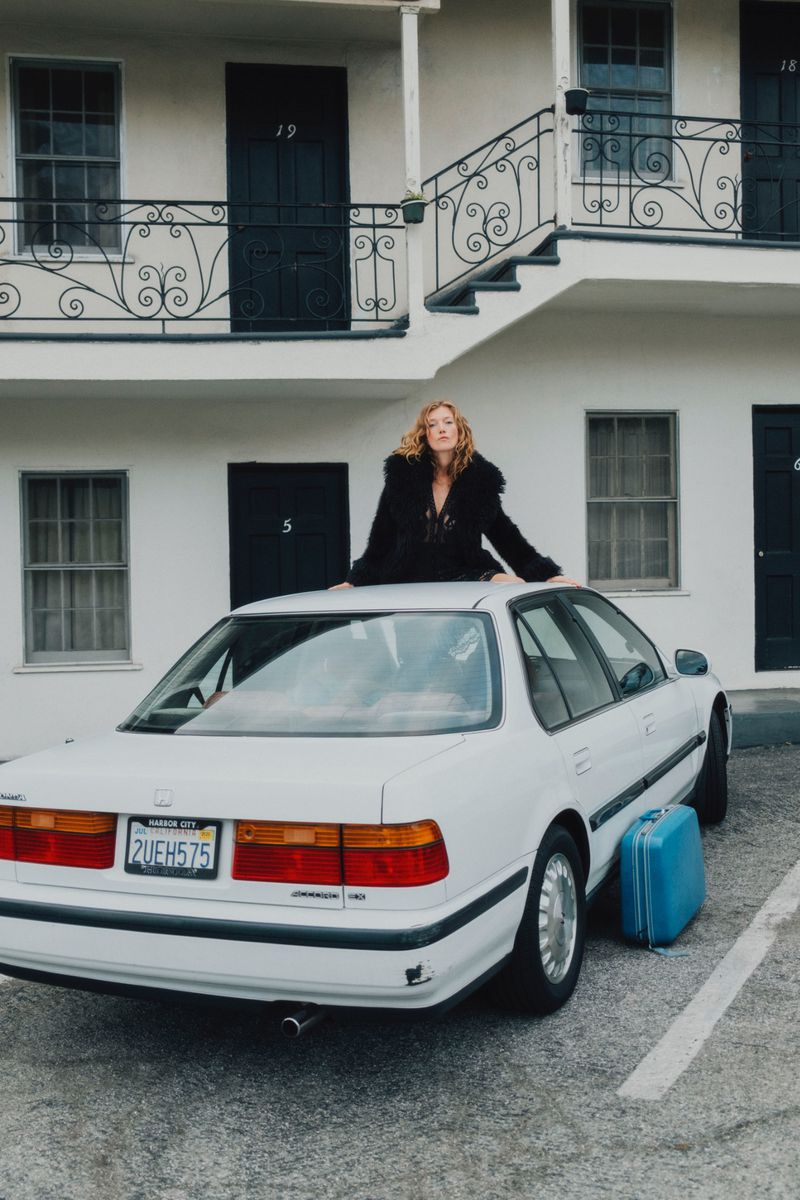 Blonde woman in black jacket poses on a white car outside an apartment motel with a blue suitcase alongside the vehicle.