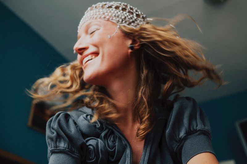 Smiling woman with curly hair wearing a beaded headpiece and a grey jacket in an indoor setting with blue wall in the background.