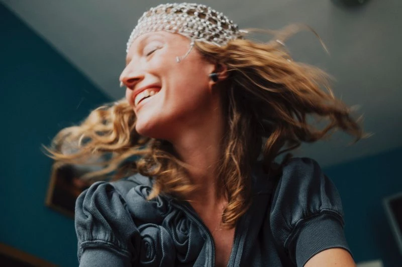 Smiling woman with curly hair wearing a beaded headpiece and a grey jacket in an indoor setting with blue wall in the background.