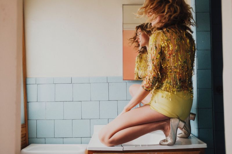 Woman in yellow shorts and a floral sequin top poses on a sink in front of a mirror in a bathroom with blue tiled walls.