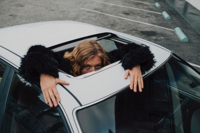 Woman in black furry coat looks out of a car sunroof in a parking lot on an overcast day, visible from chest up, cool color grading.