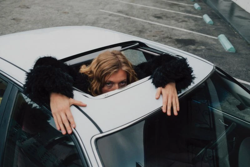 Woman in black furry coat looks out of a car sunroof in a parking lot on an overcast day, visible from chest up, cool color grading.