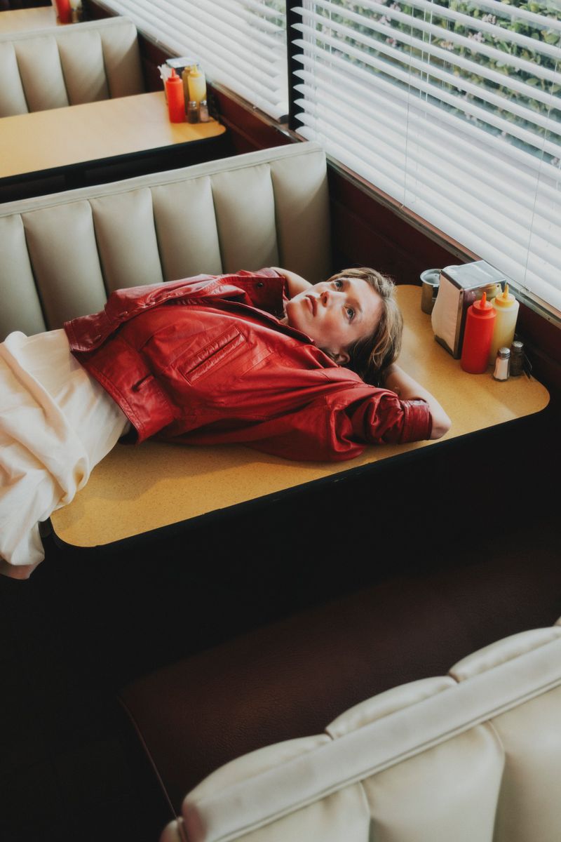 Blonde adult woman laying on a booth table wearing a red leather jacket in a diner by the window with condiments on the table.