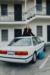Model Lou Schoof resting on the roof of a white vintage sedan with a blue suitcase parked outside a two-story motel.