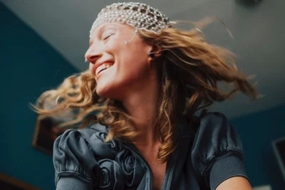 Close-up portrait of a smiling woman with tousled hair wearing a beaded headpiece and grey textured top against a dark blue wall.