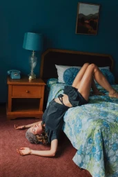 Model lying upside down on a bed with blue patterned bedding inside a dark teal bedroom.