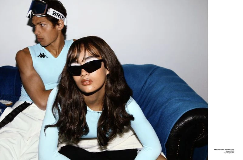 Two fashion models pose on a blue couch in a studio wearing ski goggles and sunglasses. One is wearing a light blue tank top.